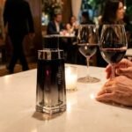 Frasco do perfume Malbec Black Legend sobre mesa branca em jantar romântico à noite com taças de vinho tinto. Cenário de restaurante de luxo com homem sofisticado e garçom ao fundo, ilustrando os melhores perfumes masculinos amadeirados para a noite.