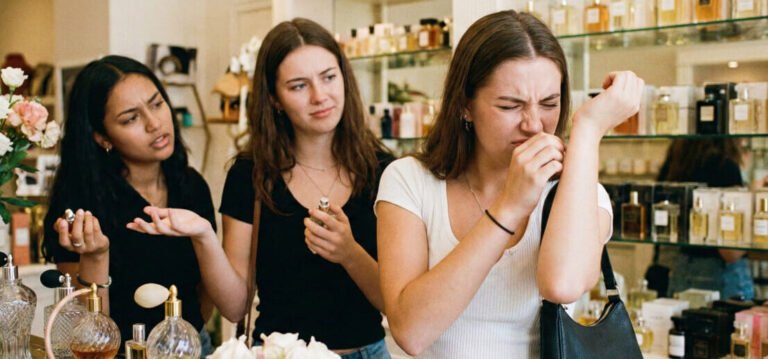 Uma jovem mulher com expressão de nojo e desgosto cheira o perfume em seu pulso dentro de uma boutique de fragrâncias, enquanto suas duas amigas a observam com expressões de confusão e surpresa.