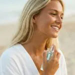 Mulher jovem, loira e bronzeada, sorrindo na praia enquanto aplica um perfume de frasco transparente no pescoço, vestindo uma saída de praia branca de linho.