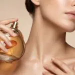 Mulher de pele iluminada aplicando perfume no pescoço, ilustrando a busca por fragrâncias de alta fixação.