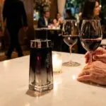 Frasco do perfume Malbec Black Legend sobre mesa branca em jantar romântico à noite com taças de vinho tinto. Cenário de restaurante de luxo com homem sofisticado e garçom ao fundo, ilustrando os melhores perfumes masculinos amadeirados para a noite.