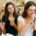 Uma jovem mulher com expressão de nojo e desgosto cheira o perfume em seu pulso dentro de uma boutique de fragrâncias, enquanto suas duas amigas a observam com expressões de confusão e surpresa.